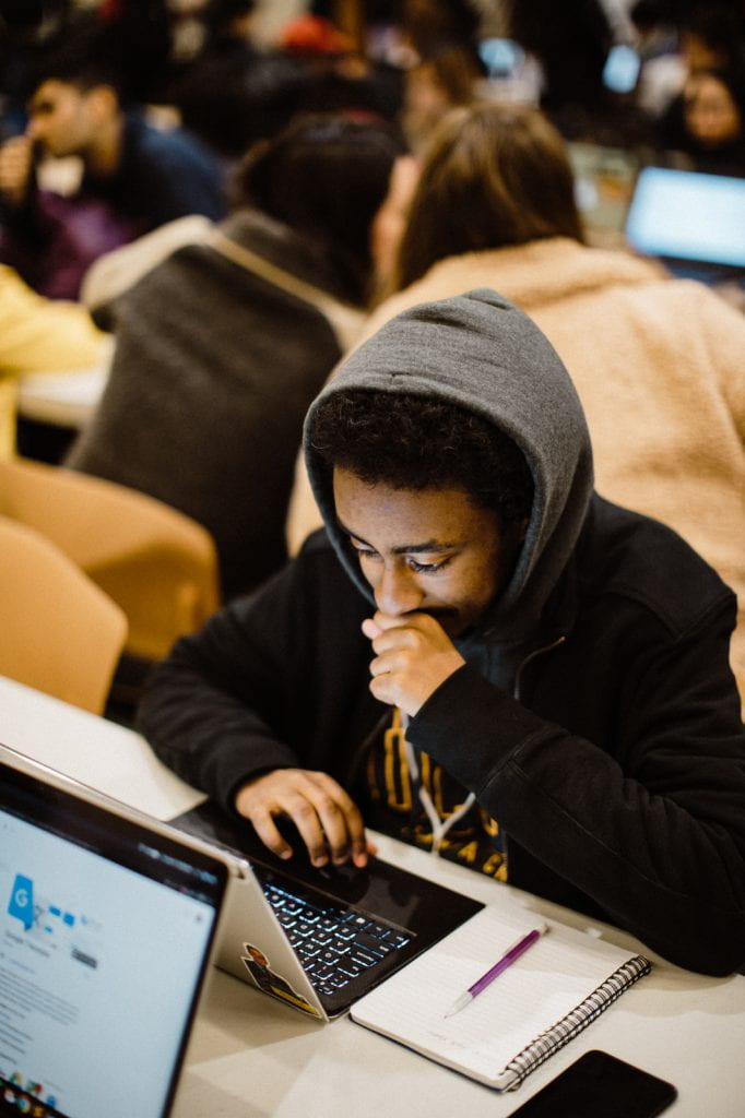 Young man with hoodie working on his laptop in a busy room.