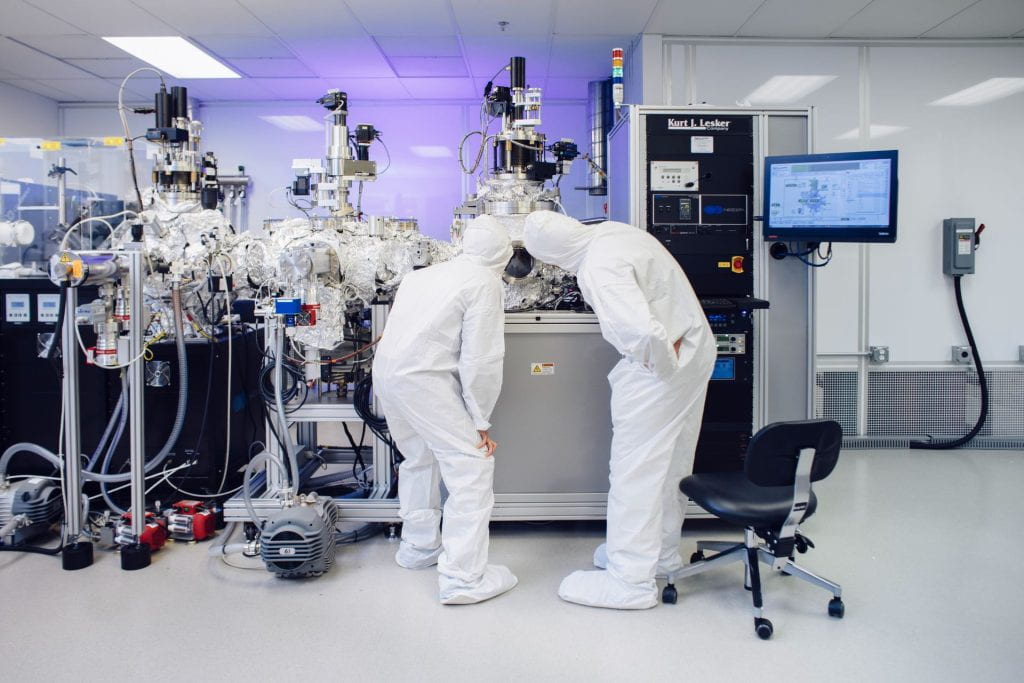 Two people working in a research laboratory environment, wearing full body suits and surrounded by lab equipment. 